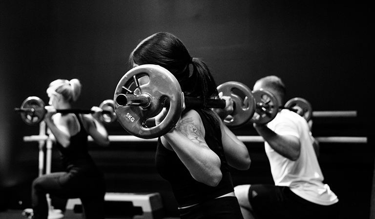 Black and white photo of woman doing single-leg, shoulder supported weighted squats alongside a class of others.