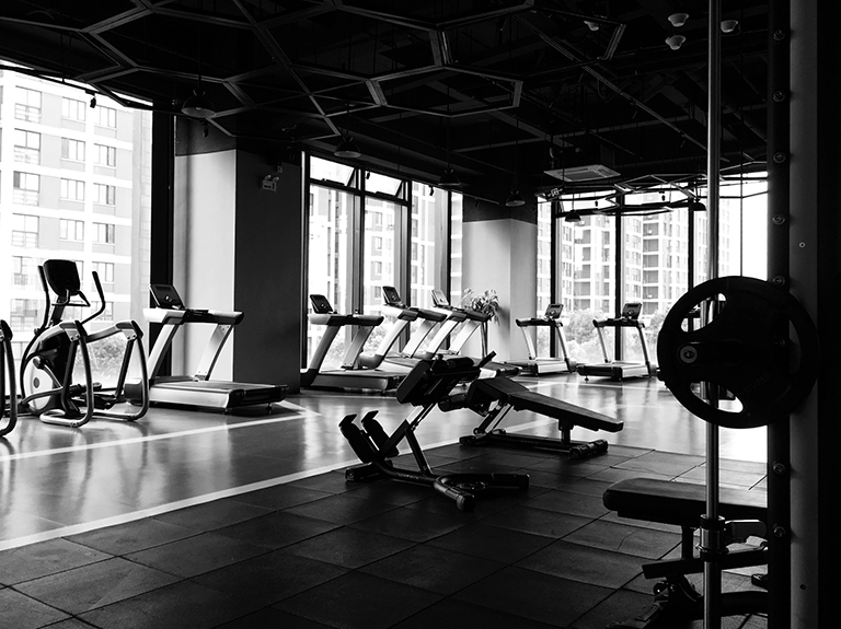 Black and white photo of gym. Treadmills and cardio equipment line windows in a upper floor gym scene in the background, with padded flooring and weightlifting equipment in the foreground.